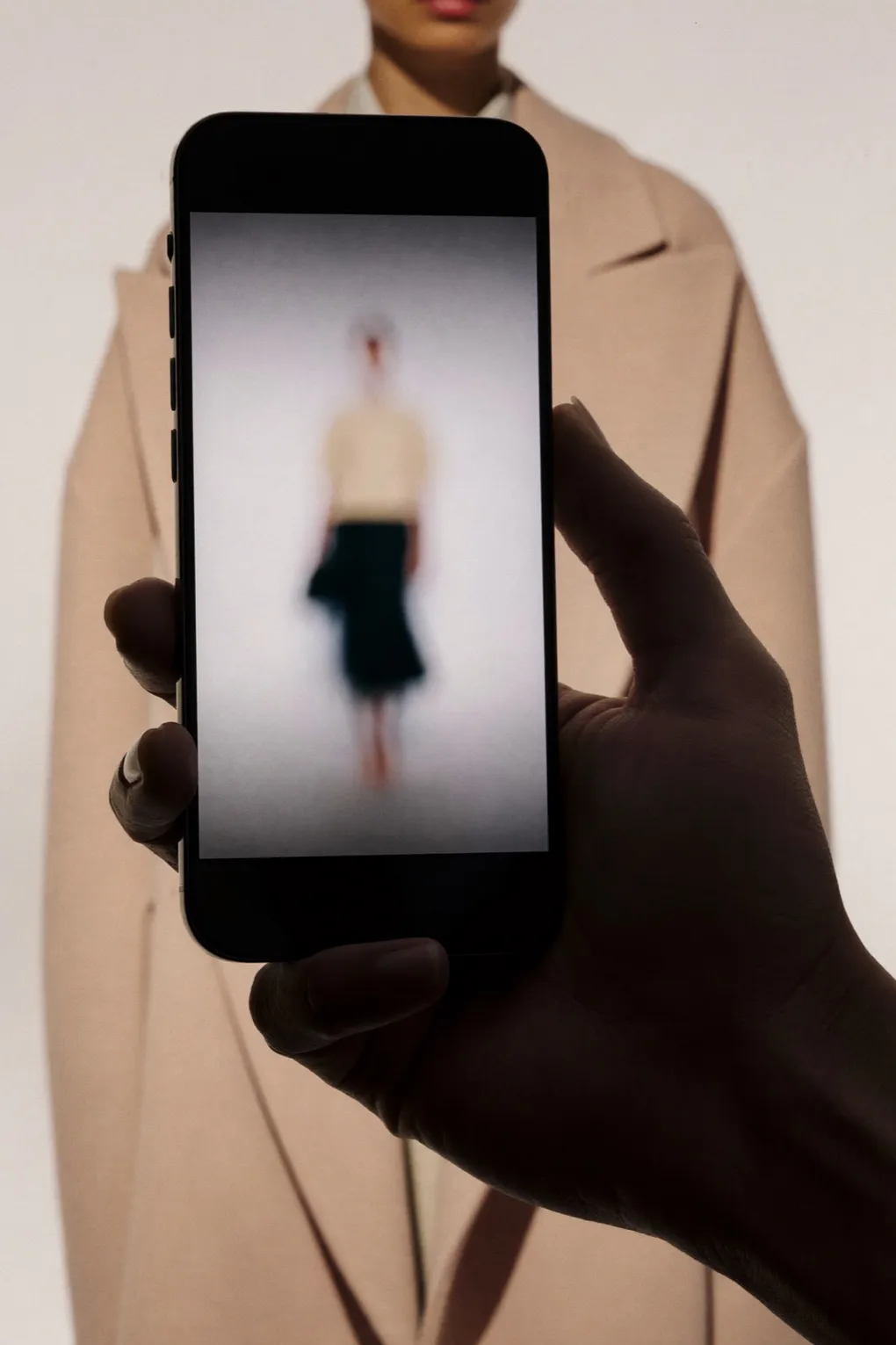 Hand hält ein Smartphone mit einer unscharfen Modelaufnahme vor einen Kamelmantel.