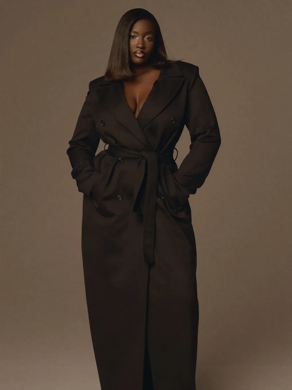 Curve Model im dunklen Trenchcoat vor braunem Studiohintergrund.