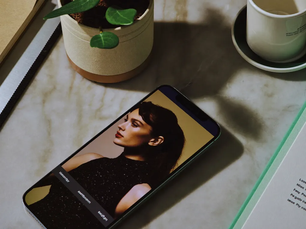 Smartphone auf einer Marmorplatte zeigt ein Modelporträt, daneben Pflanze und Tasse.