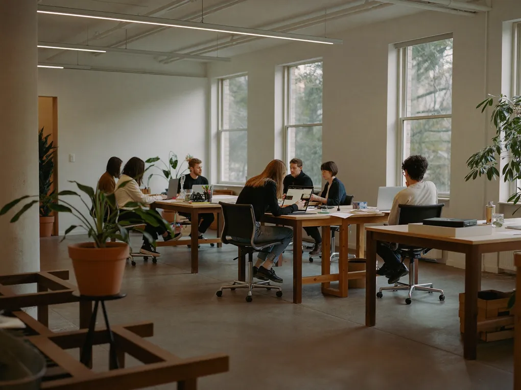 Großraumbüro mit langen Holztischen, an denen mehrere Personen arbeiten.