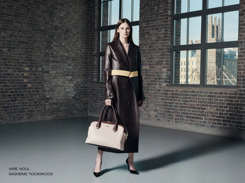 Model mit Tasche in einem Loft mit hohen Sprossenfenstern und Backsteinwand.