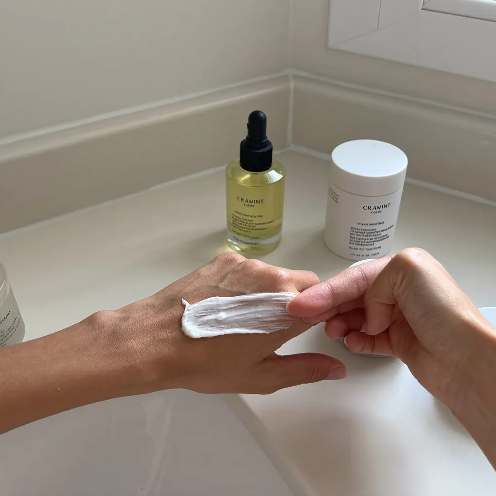 Hände tragen Creme auf, dahinter ein Serum und ein Tiegel auf hellem Untergrund.