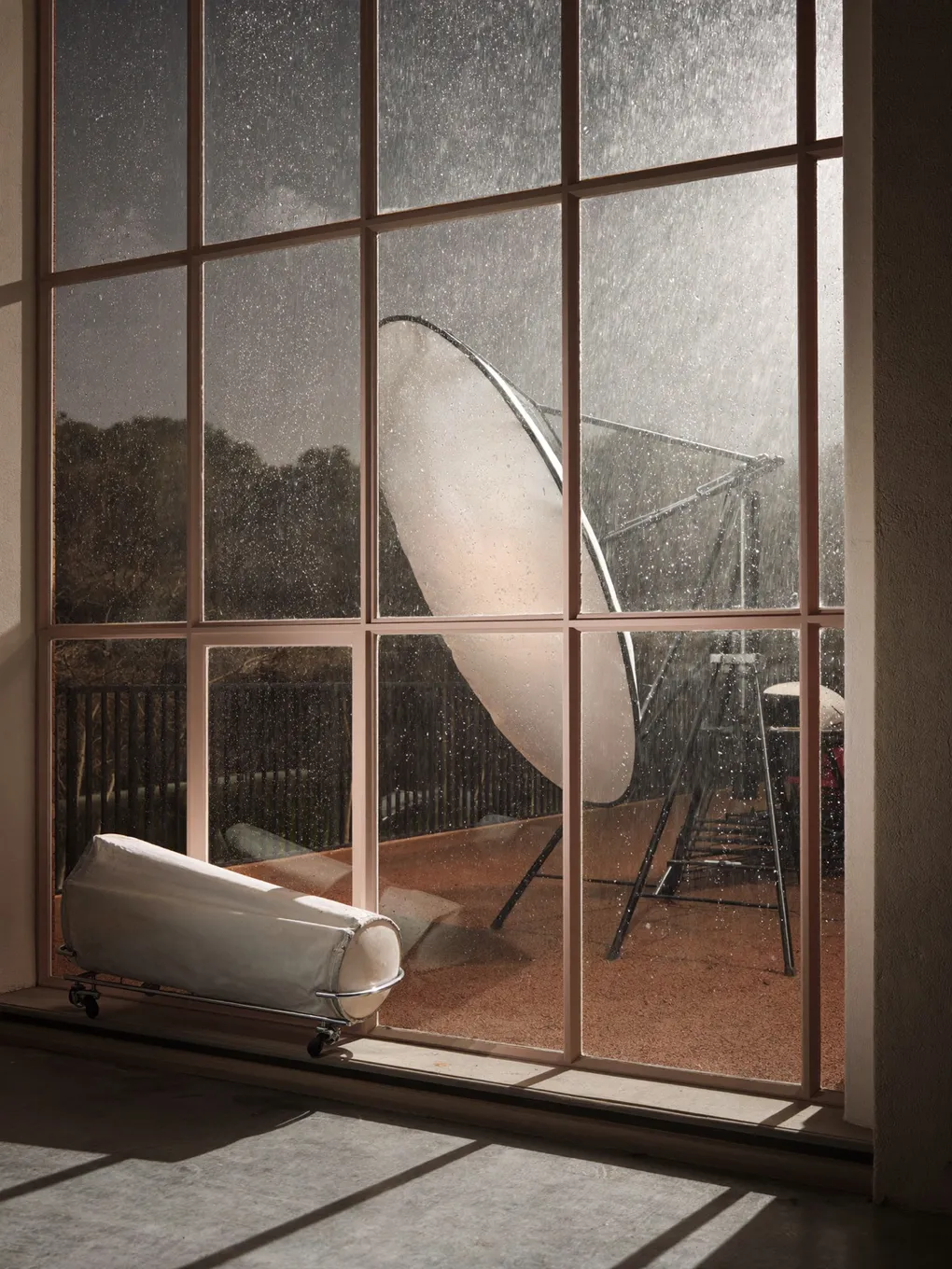 Blick durch ein großes Fenster auf eine Terrasse mit Reflektor bei bedecktem Himmel.